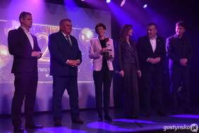 Pokaż zdjęcie: Grupa sześciu osób w eleganckich strojach stoi na scenie oświetlonej fioletowym światłem. W tle widać świetlne efekty oraz ekran, a jedna osoba trzyma w ręku różę. Wszystkie osoby są ubrane elegancko.