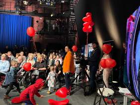 Pokaż zdjęcie: Zdjęcie studia telewizyjnego, goście siedzą na krzesłach, obok stoi Pani redaktor Anna Dąbrowska w żółtej marynarce oraz kamerzysta.