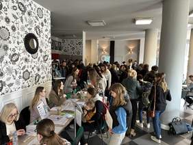 Pokaż zdjęcie: Na Sali z filarami i wzorzystą tapetą na ścianie stoi dużo ludzi. Po lewej długi stół z napisem powiatowy Urząd Pracy na którym leży duża ilość materiałów. Za nim kilka kobiet w marynarkach udziela informacji.