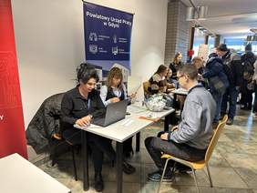 Pokaż zdjęcie: Za biurkiem siedzi kobieta w jasnej marynarce i okularach. Przy stoliku Napis FAZON oraz dwa banery jedne z napisem FAZON drugi z logo ściskających się dłoni.  Naprzeciw siedzi jasnowłosa kobieta w jasnoszarej kurtce z przewieszonym szalem w kratkę 