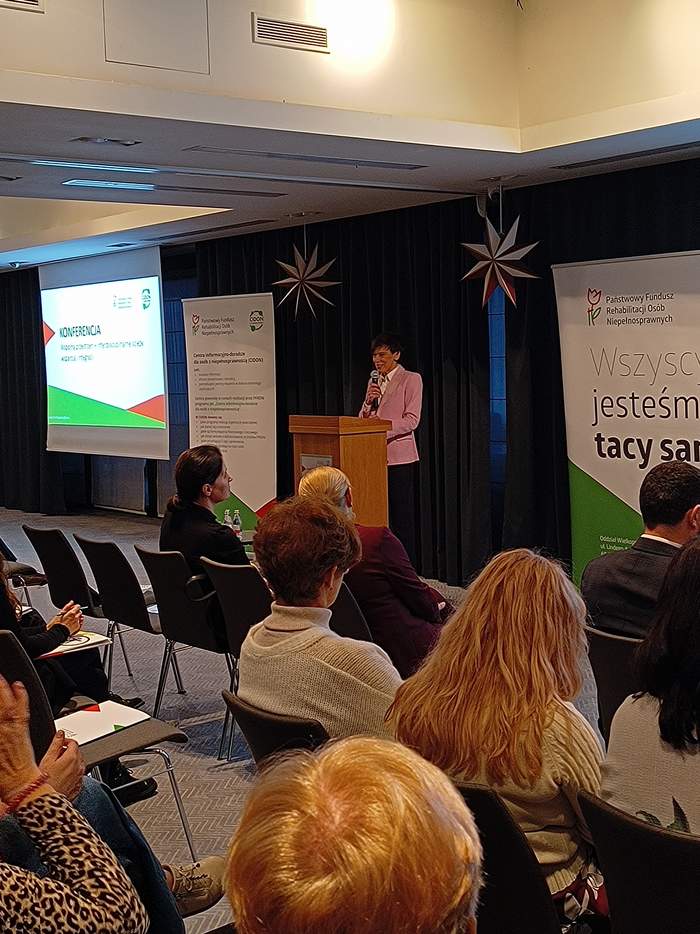 Pokaż zdjęcie: Na zdjęciu widać grupę osób siedzących na krzesłach, zwróconych twarzami w stronę mównicy. Przy mównicy stoi kobieta w różowej marynarce, przemawia do mikrofonu. Na scenie ustawione są dwa banery; jeden z napisem „Państwowy Fundusz Rehabilitacji Osób Niepełnosprawnych”, drugi z fragmentem hasła „Wszyscy jesteśmy tacy sami”. Po lewej stronie sali znajduje się ekran z rzutnikiem z napisem „Konferencja”.