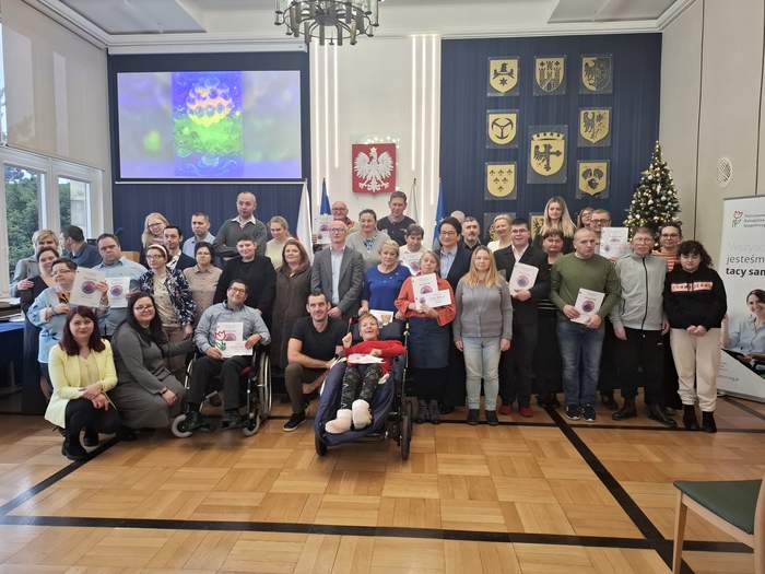 Pokaż zdjęcie: Grupa osób pozuje do fotografii. Uczestnicy trzymają w rękach otrzymane dyplomy i nagrody. Osoby z niepełnosprawnościami, w tym na wózkach, stoją i siedzą w pierwszym rzędzie