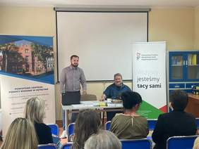 Pokaż zdjęcie: Dwóch mężczyzn siedzi przy biurku, na którym znajdują się ulotki. Koło nich znajduje się baner PFRON i PCPR. Przed nimi kilka osób siedzących na krzesłach.