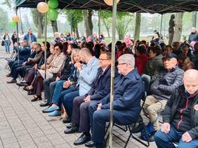 Pokaż zdjęcie: Na krzesłach siedzą ludzie. Oglądają występy artystyczne odbywające się na scenie. W tle widać park miejski. 