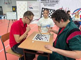 Przy stole siedzi dwóch zawodników Turnieju w Warcaby Uczestników Warsztatów Terapii Zajęciowej. Rozgrywają patrię warcabów. Obok nich siedzi dziewczyna, która jest sędzią pojedynku. 