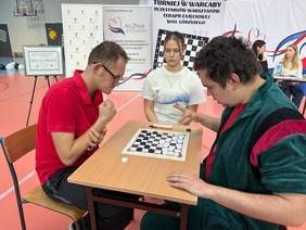 Pokaż zdjęcie: Przy stole siedzi dwóch zawodników Turnieju w Warcaby Uczestników Warsztatów Terapii Zajęciowej. Rozgrywają patrię warcabów. Obok nich siedzi dziewczyna, która jest sędzią pojedynku. 