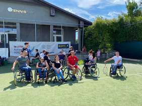 Pokaż zdjęcie: Na tle budynku Klubu tenisowego jest ustawionych kilkanaście osób na wózkach inwalidzkich, które pozują do zdjęcia. Część z nich trzyma w rękach rakiety tenisowe.