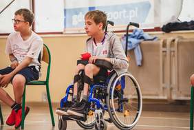 Pokaż zdjęcie: Na Sali gimnastycznej chłopiec siedzący na wózku inwalidzkim trzyma w ręku piłkę i patrzy w jakiś cel. Obok siedzi chłopiec na krześle.