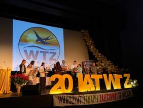 Na scenie podczas jubileuszu 20-lecia Warsztatu Terapii Zajęciowej w Wałczu stoi grupa uczestników i przedstawicieli zaproszonych gości. W centralnej części znajduje się duża dekoracja z napisem „20 lat WTZ”, a w tle widoczne jest logo warsztatu przedstawiające lecącą jaskółkę. Scenę zdobią czerwone róże oraz kompozycja balonów w kolorach złotym, srebrnym i białym.