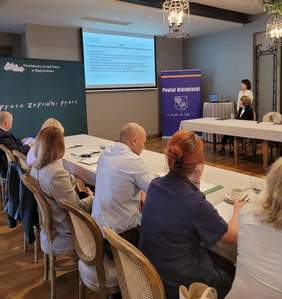 Pokaż zdjęcie: Po prawej stronie sali dwie kobiety jedna siedzi druga stoi, omawia slajd który  wyświetla się na ekranie. Po lewej stronie przy stołach siedzą kobiety i mężczyźni na stołach leżą notatki.