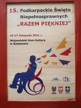 Pamiątkowa tabliczka drewniana z napisem 15 Podkarpackie święto niepełnosprawnych, daty poniżej osoba na wózku odlatują od niej jakby kolorowe kartki
