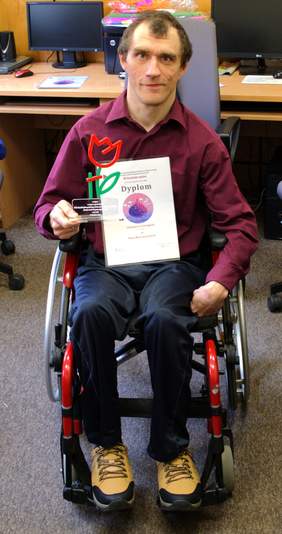 Pokaż zdjęcie: Mężczyzna o krótkich, ciemnych włosach siedzi na wózku inwalidzkim o czerwonej ramie. Trzyma dyplom oraz statuetkę w kształcie czerwonego tulipana na zielonej łodydze. Ubrany jest w bordową koszulę, ciemne spodnie i brązowo-żółte buty trekkingowe.