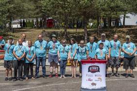 Pokaż zdjęcie: Grupa uczestników Wojewódzkiej Spartakiady Osób Niepełnosprawnych 2024 w Ogrodniczkach pozuje do zdjęcia z medalami, stojąc przed lasem i stoiskiem organizatorów.