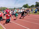 Po czerwonej bieżni biegnie kilku sportowców z niepełnosprawnością. W tle widać także innych zawodników. 