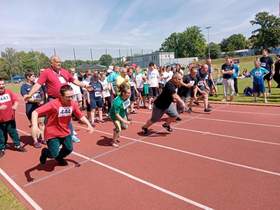 Po czerwonej bieżni biegnie kilku sportowców z niepełnosprawnością. W tle widać także innych zawodników. 