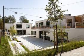 Pokaż zdjęcie: Dwa białe, prostokątne budynki piętowe, z dużymi przeszkleniami w budynku po prawej duży taras. Przed nimi bujna trawa i drzewa. Nad nimi sieć lamp.