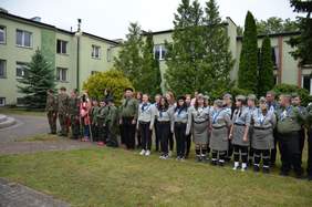 Pokaż zdjęcie: Zbiórka harcerzy. Stoją w dwuszeregu. Wśród nich dzieci i dorośli. W tle budynek i drzewa. 