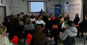 Pokaż zdjęcie: Grupa około trzydziestu osób znajduje się w sali konferencyjnej i przysłuchuje się wystąpieniu osoby prowadzącej szkolenie. W tle duży ekran z napisem: „Agenda”. Po obu stronach ekranu ustawione są banery organizatorów spotkania.