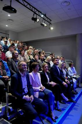 Pokaż zdjęcie: Grupa kilkudziesięciu osób siedzi w dużej sali, w której krzesła usytuowane są w układzie kinowym. Pod sufitem znajdują się reflektory doświetlające salę.