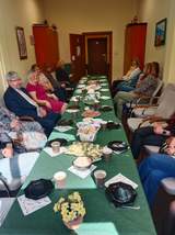 Przy podłużnym stole siedzą ludzie. Trwa Wielkanocne Śniadanie. Na stole leżą różne potrawy i dekoracje świąteczne. 