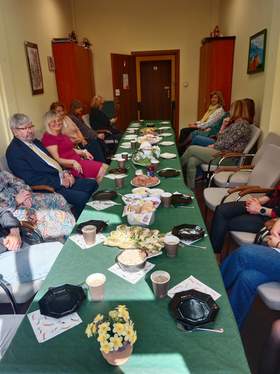 Przy podłużnym stole siedzą ludzie. Trwa Wielkanocne Śniadanie. Na stole leżą różne potrawy i dekoracje świąteczne. 