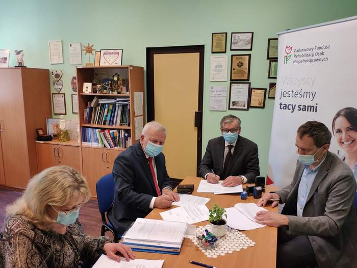 Pokaż zdjęcie: Przy stole siedzą 4 osoby, 3 mężczyzn podpisuje dokumenty, kobieta patrzy na dokument przed sobą