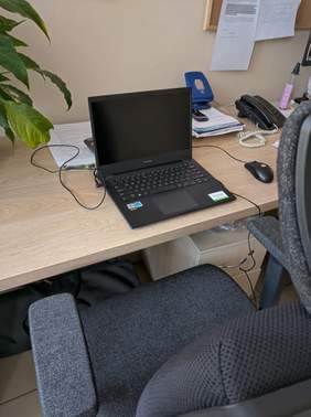 Pokaż zdjęcie: Fragment biurka na którym znajduje się laptop, dokumenty i telefon, na pierwszym planie fragment czarnego fotela