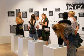 Pokaż zdjęcie: Fragment pomieszczenia, 6 sób ogląda białą ścianę z czarnymi tablicami zapisanymi białym tekstem. Jedna osoba pochyla się na białą gablotą