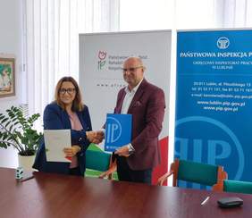 Pokaż zdjęcie: Za stołem konferencyjnym kobieta i mężczyzna pozują do zdjęcia trzymając w rękach teczki z podpisanymi umowami.