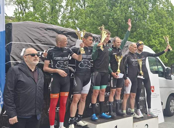 Pokaż zdjęcie: Zawodnicy stoją na podium podczas dekoracji zwycięzców wyścigu kolarskiego, trzymają puchary i unoszą je w górę. Obok znajdują się organizatorzy i kibice, w tle widoczny samochód, banery oraz drzewa.