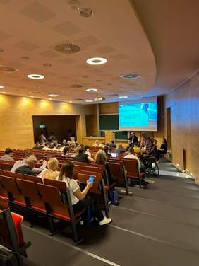 Pokaż zdjęcie: Zdjęcie z tyłu sali wykładowej w kierunku prelegenta, na projektorze wyświetlany jest slajd z prezentacji.