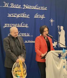 Pokaż zdjęcie: Przy mównicy stoją dwie osoby, kobieta w czerwonej marynarce, mężczyzna z wąsami i ciemnej marynarce trzyma w ręku koszyk z cukierkami.