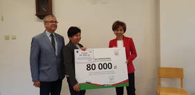 Pokaż zdjęcie: trzy osoby stoją obok siebie. Dwie kobiety trzymają dużą tablicę symbolizującą czek. 