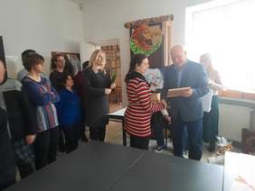 Pokaż zdjęcie: Zdjęcie w sali artystycznej WTZ w Hrubieszowie. Na zdjęciu ok. 12 osób. Mężczyzna przekazuje kobiecie dyplom i uścisk dłoni.