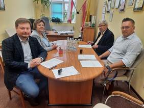 Pokaż zdjęcie: Przy podłużnym stole siedzą cztery osoby: kobieta i trzech mężczyzn. Wszyscy patrzą w stronę obiektywu aparatu. Na stole leżą dokumenty, pieczątki, długopisy i woda mineralna. W tle wiszą flagi: Polski i Łodzi. 