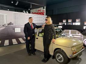 Pokaż zdjęcie: Mężczyzna ubrany w granatowy garnitur białą koszulę i czerwony krawat rozmawia z kobietą ubraną na czarno w okularach przeciwsłonecznych. Obok nich stoi samochód Marki Fiat Maluch. Za nimi zdjęcie bloków z wielkiej płyty obok napis Solidarnoś