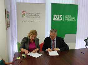 Pokaż zdjęcie: Przy stole siedzi kobieta blondynka obok niej mężczyzna w garniturze, na stole leżą dokumenty do podpisania. W tle widać rollupy z logo PFRON oraz ZUS.za ich