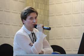 Pokaż zdjęcie: Kobieta blondynka z krótkimi włosami trzyma przy ustach mikron, siedzi za stołem konferencyjnym