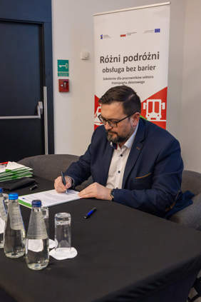 Pokaż zdjęcie: Pan Patryk Świrski Prezes Zarządu Kolei Śląskich siedzi przy stole i podpisuje deklarację dotyczącą Partnerstwa na rzecz dostępnej obsługi różnych podróżnych. W tle baner z napisem Różni podróż – obsługa bez barier.