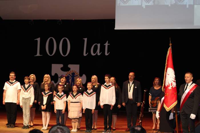 Pokaż zdjęcie: Na scenie znajduje się grupa uczniów za nimi stoją nauczyciele.Z prawej strony sceny mężczyzna stoi ze sztandarem. Wszyscy śpiewają