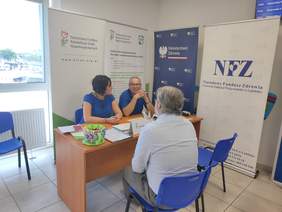 Pokaż zdjęcie: Przy stoliku informacyjny na którym leżą ulotki, siedzą trzy osoby, kobieta i dwóch mężczyzn, rozmawiają.