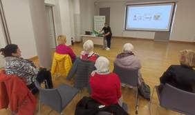 Pokaż zdjęcie: Grupa seniorów siedzi na krzesłach w sali konferencyjnej i słucha prezentacji prowadzonej przez kobietę stojącą przy stole. Na ekranie w tle wyświetlana jest prezentacja multimedialna dotycząca technologii wspomagających.