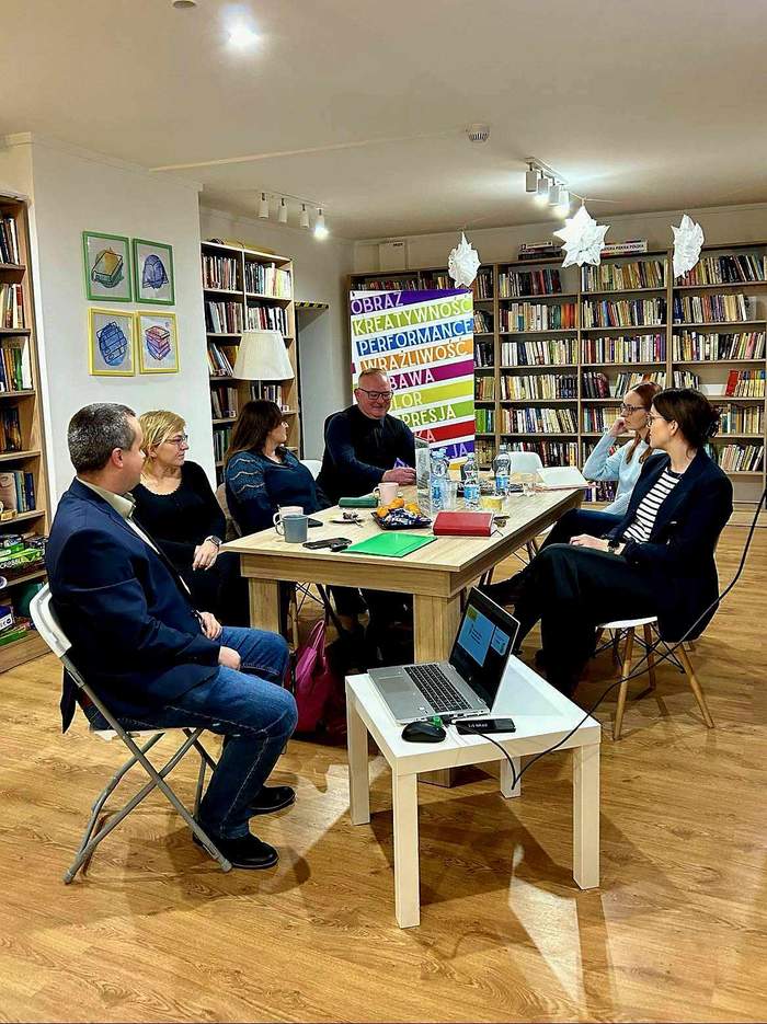 Pokaż zdjęcie: W bibliotece w której stoją regały z książkami, siedzą przy stoliku goście spotkania i prowadzą rozmowę. Jest widoczny laptop na stoliku i inne materiały informacyjne przy stole, a także roll-up biblioteki