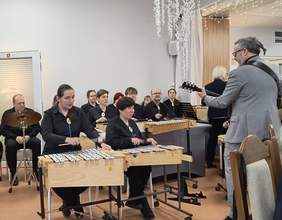 Pokaż zdjęcie:  Na tym zdjęciu widoczna jest grupa muzyków grających na ksylofonach i innych instrumentach. Do grupy dołączył mężczyzna w eleganckim garniturze, grający na gitarze. Występ odbywa się w ozdobionej sali, z widocznymi dekoracjami świątecznymi i lampkami na suficie. Wszyscy uczestnicy występu wyglądają na skupionych, a atmosfera wydaje się ciepła i uroczysta.