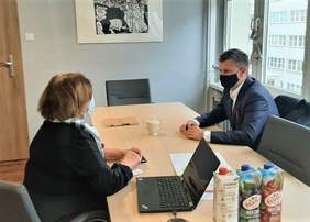 Pokaż zdjęcie: Tyłem siedzi Dyrektor Oddziału Aleksandra Gierej, ubrana w czarną sukienkę i jasny szal, przed nią, na stole leży otwarty laptop. Przed nią siedzi Pan Michał Ramlau - Wicestarosta Powiatu Toruńskiego, ubrany w ciemny garnitur i białą koszulę, oboje są w maseczkach.