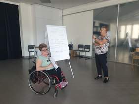 Na zdjęciu widzimy dwie uczestniczki warsztatów. Jedna z nich siedzi na wózku inwalidzkim i trzyma kartkę, podczas gdy druga osoba stoi przy flipcharcie z zapisanymi notatkami. Prowadzą one dyskusję na temat dostępności, a zapisane na tablicy punkty dotyczą wniosku o dostępność. 