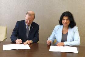 Kobieta i mężczyzna siedzą przy drewnianym stole. Podpisują dokumenty - porozumienie. Oboje są elegancko ubrani.