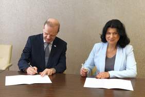 Pokaż zdjęcie: Kobieta i mężczyzna siedzą przy drewnianym stole. Podpisują dokumenty - porozumienie. Oboje są elegancko ubrani.