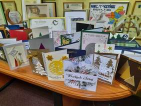 Pokaż zdjęcie: Na stoliku leży mnóstwo nadesłanych kartek z życzeniami bożonarodzeniowymi. W tle widać także różne dyplomy i podziękowania. 