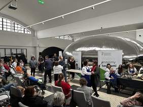 Pokaż zdjęcie: Duża przestrzeń, na środku tańczą trzy pary. Obok krzesła na których siedzą goście po prawej stronie uczestnicy zespołu siedzą przy stole. Po lewej stronie gra drugi zespół na gitarach i klawiszach. W tle banery z logo PFRON z napisem wszyscy